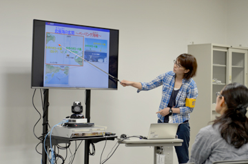 モニターを指し示して説明する女性と話を聞く女性