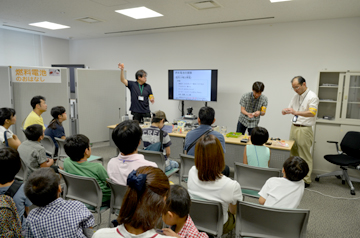 前方で説明する3人のスタッフと、席で話を聞く見学者たち