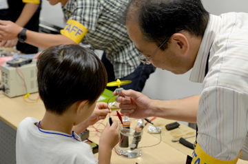 ビーカーの燃料電池を示しながら、子どもに電極の働きを説明する研究者