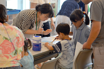浅い容器に筆で絵を描く子どもと、それを説明する女性