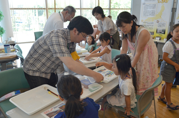 スタッフが手順を示し、子どもと大人が砂絵の体験している様子