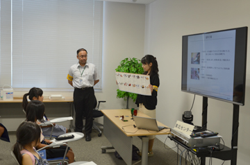 講義室でスタッフが資料を掲げて説明し、子どもたちが聞いている様子