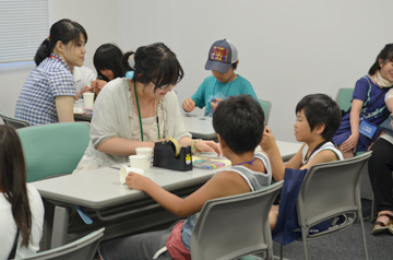 机を囲んで工作に取り組む子どもたちと、それをサポートする大人