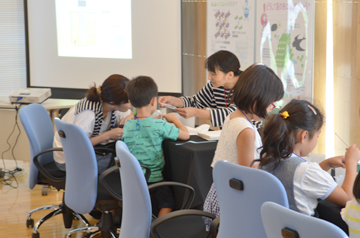 テーブルでスタッフと向かい合って子どもたちが説明を受けながら工作に取り組む様子