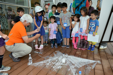 バルコニーでスタッフが子どもたちに実験の様子を見せている場面