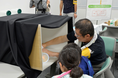 スタッフが黒い箱の中の実験装置を子どもに説明している様子