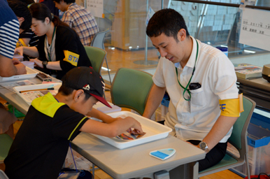 トレイに並んだ教材を前に、スタッフが子どもたちへ作業手順を説明している様子