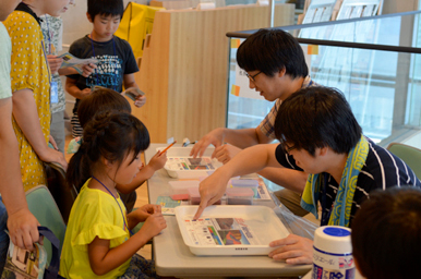 トレイに並んだ教材を前に、スタッフが子どもたちへ作業手順を説明している様子
