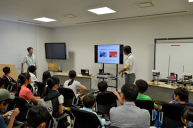 モニターに映る資料を前に、スタッフが実験装置の仕組みを参加者に説明している様子