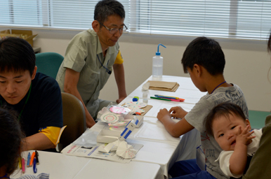 ろ紙やペンの教材を前に、スタッフが子どもに工作の手順を説明している様子