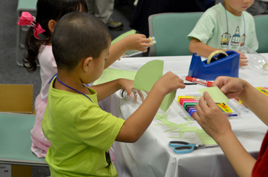 テーブルで子どもが色画用紙を使って工作に取り組み、大人がそばで手助けしている様子