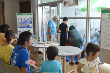 室内で子どもたちが座って展示を見学し、奥ではスタッフが親子に説明している様子