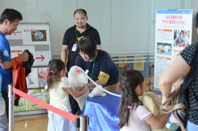 スタッフがロボットのパロを子どもに手渡し、周囲の大人たちが興味深く見ている様子