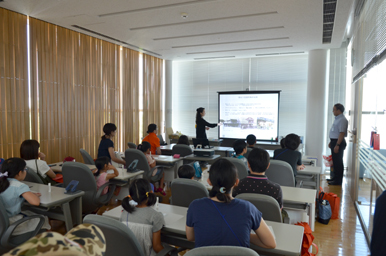 講義室でスクリーンの説明を聞いている参加者たちと、前方で発表するスタッフ