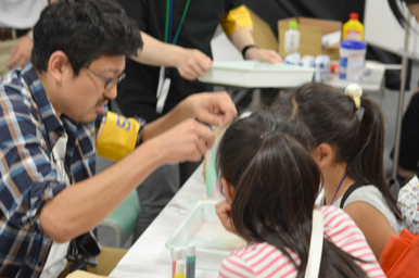 スタッフが透明な容器の前で実験の手順を示し、子どもたちが熱心に見守っている様子