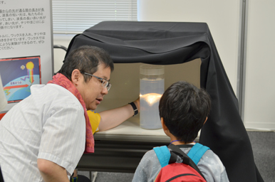 暗幕で囲われた装置の中を指さしながら、円筒形の容器に光が当たる様子を子どもに説明している場面