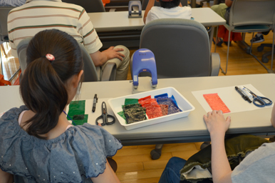 机の上に並んだ色付きフィルムやハサミなどの材料を前に、参加者が作業の準備をしている様子