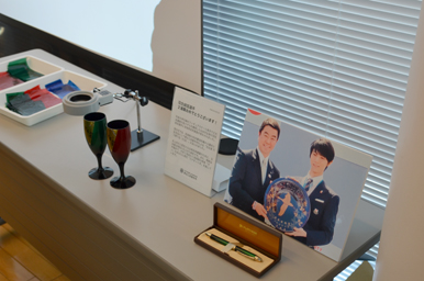 玉虫塗のワインカップやボールペンと、県民栄誉賞の大皿贈呈を紹介するパネルが並ぶ、玉虫塗を紹介する展示テーブル