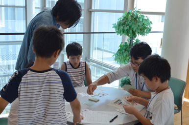 丸いテーブルを囲んで、子どもたちがカードゲーム取り組み、大人がそばで見守っている様子