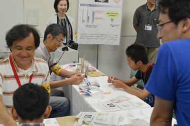 実験素材を使いながら、スタッフが子どもに手順を説明し、周囲の来場者が見守っている様子