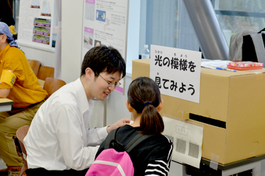 『光の模様を見てみよう』と書かれた装置を前に、スタッフが子どもに説明している様子真
