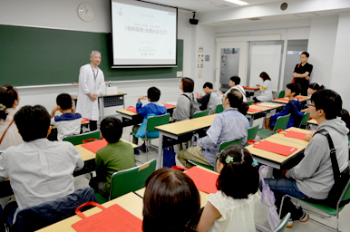教室で講師が前方に立って話をし、参加者が机に着席して説明を聞いている様子真