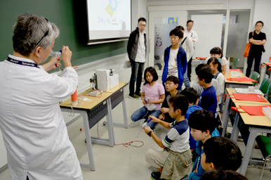 講師が実験器具を示しながら説明し、子どもたちが前に集まって話を聞いている様子