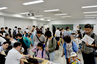 教室内の複数の体験ブースで、来場者がパソコンや展示物を使って活動し、スタッフが説明しているにぎやかな様子