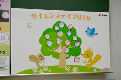 サイエンスデイ2019のボードに、リスやキツネ、鳥のイラストとともに、クロマトグラフィーで作った花のような模様の作品が木の絵に貼られて展示されている様子